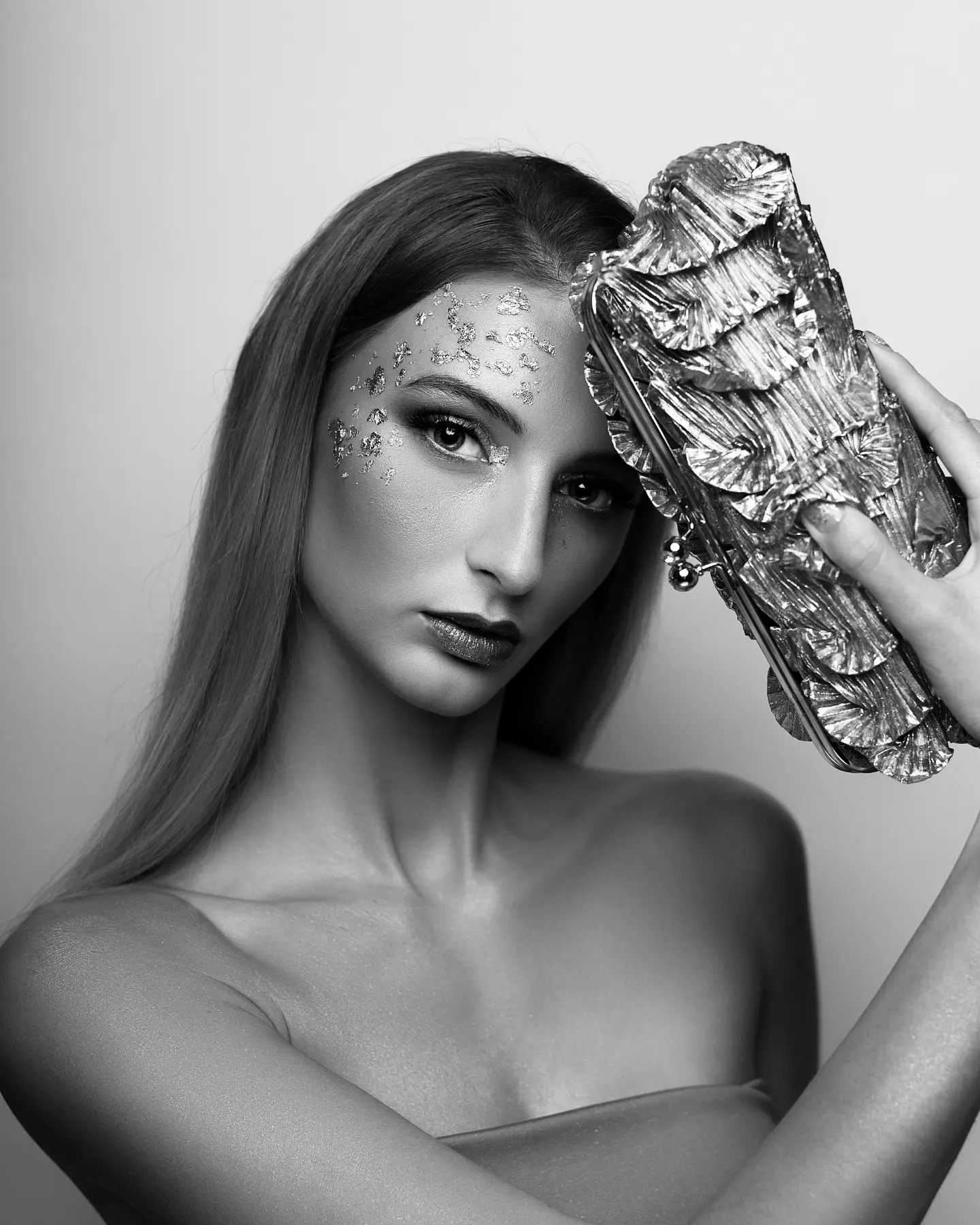 Czarno-białe portretowe ujęcie modelki z folią na twarzy i wyrazistym makijażem oczu, trzymającej srebrną, teksturowaną torebkę w Myszkowie.