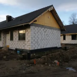 Budowa bliźniaka z białego bloczka w Jeleniej Górze, widoczne fragmenty dachu z czarnej dachówki, ocieplenie ścian szczytowych płytami OSB, teren wokół w stanie surowym.