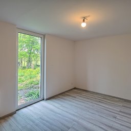 Mikołaj Janowski - Pusty pokój z nową podłogą imitującą drewno, świeżo pomalowanymi ścianami w odcieniu beżu, grzejnikiem pod oknem i pojedynczą żarówką na suficie.