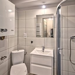 Mikołaj Janowski - Jasna łazienka z białymi kwadratowymi płytkami, kabiną prysznicową z zaokrąglonymi drzwiami, wiszącą szafką z umywalką, toaletą i piecem gazowym na ścianie.