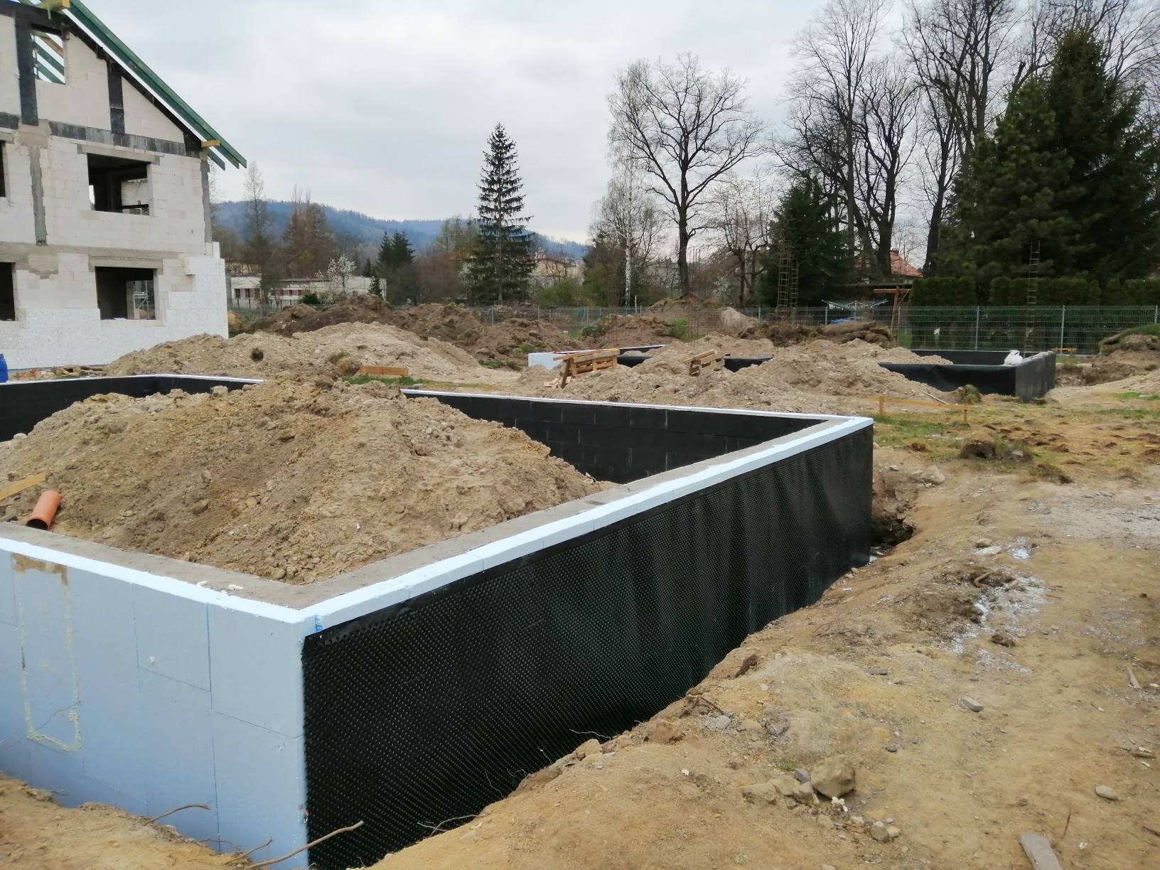 Fundament domu w trakcie budowy, ściany zabezpieczone czarną folią kubełkową i ocieplone styropianem, z widoczną stertą ziemi i fragmentem budynku w stanie surowym otwartym.
