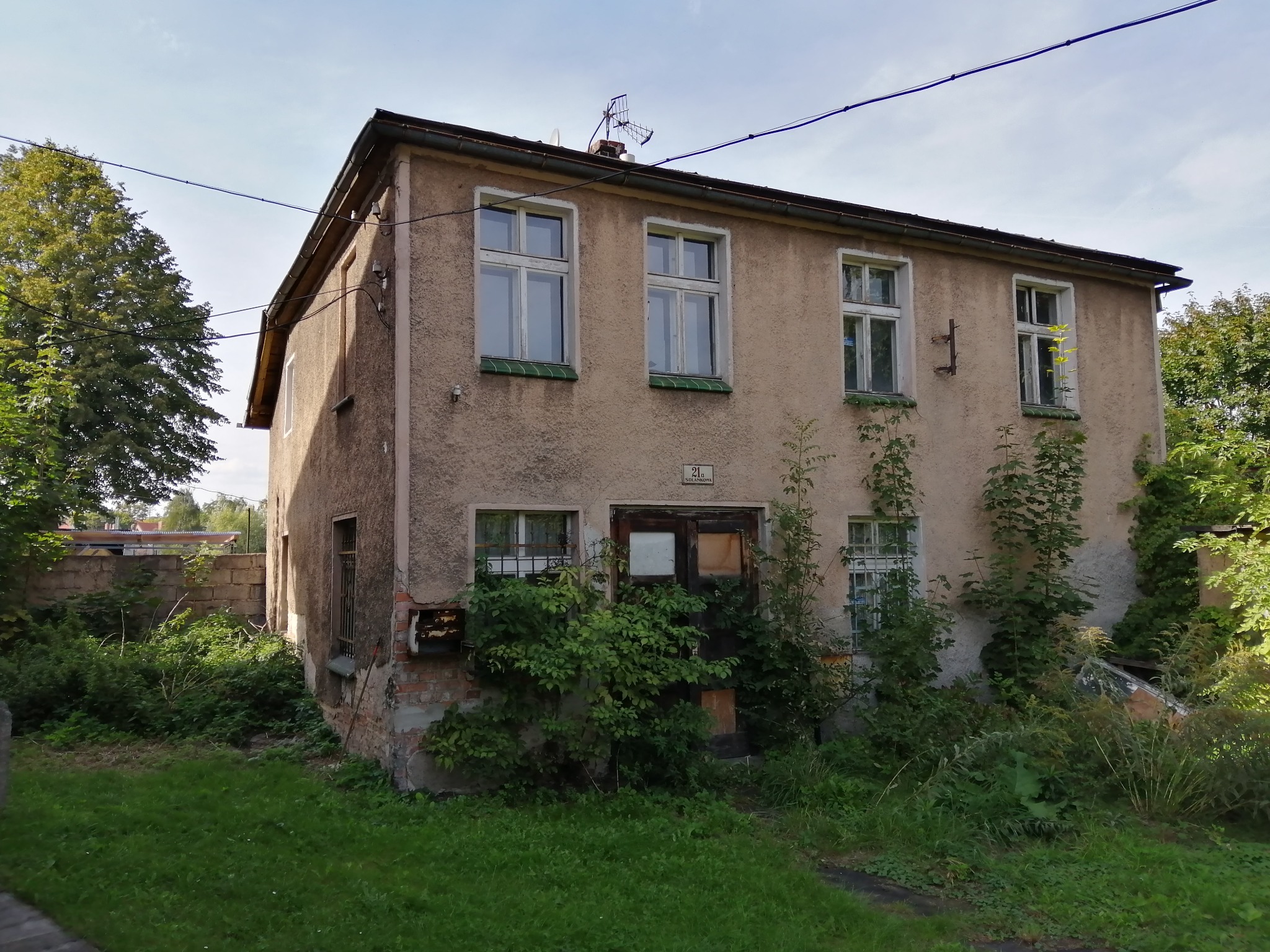 Dwupiętrowy, zaniedbany budynek z jasną elewacją i zarośniętym wejściem, widoczne ślady korozji i odpadającego tynku.