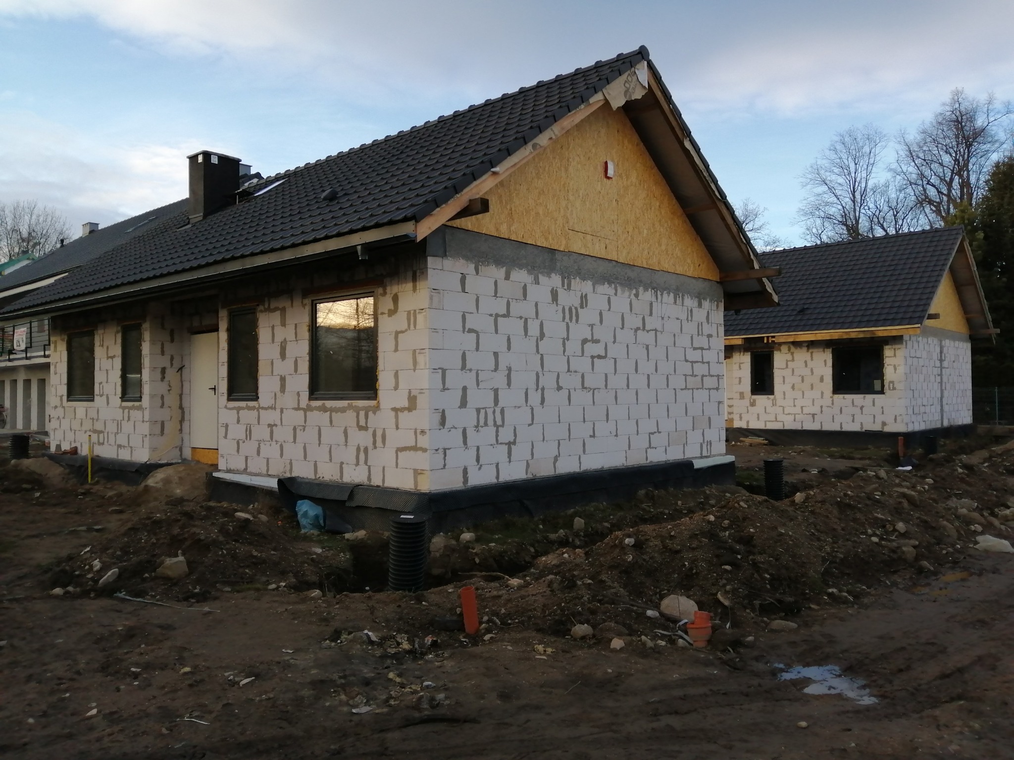 Budowa bliźniaka z białego bloczka w Jeleniej Górze, widoczne fragmenty dachu z czarnej dachówki, ocieplenie ścian szczytowych płytami OSB, teren wokół w stanie surowym.