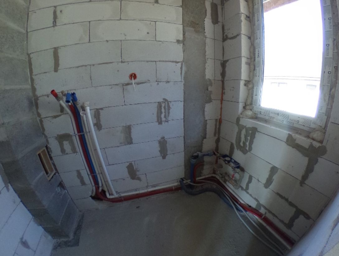 Surowa ściana z betonu komórkowego z widocznymi, nowo poprowadzonymi rurami instalacji hydraulicznej w kolorach czerwonym, niebieskim i białym, z oknem w tle.