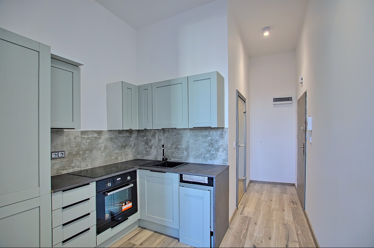 Nowoczesna, jasna kuchnia z szarymi szafkami, czarnym blatem i płytą indukcyjną, widok na przedpokój z drewnopodobną podłogą i drzwiami.