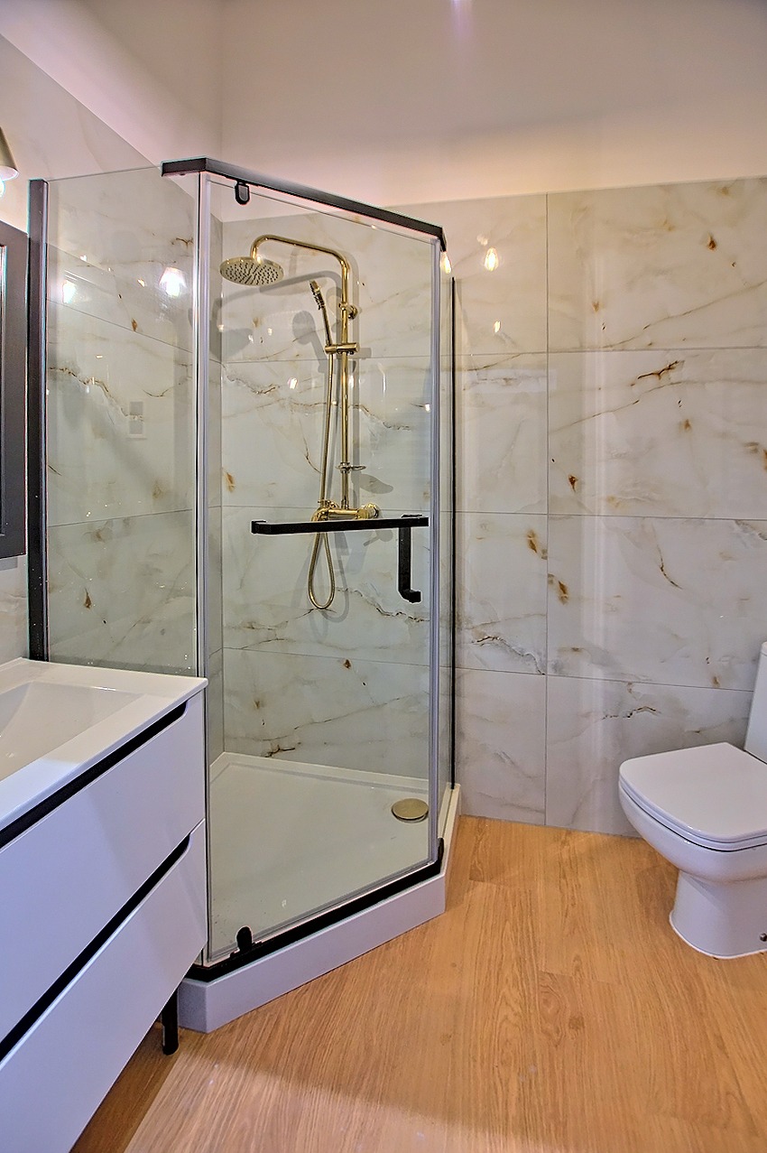 Nowoczesna łazienka z kabiną prysznicową walk-in, złotą armaturą, umywalką z szafką i toaletą, wykończona płytkami imitującymi marmur i drewnopodobną podłogą.