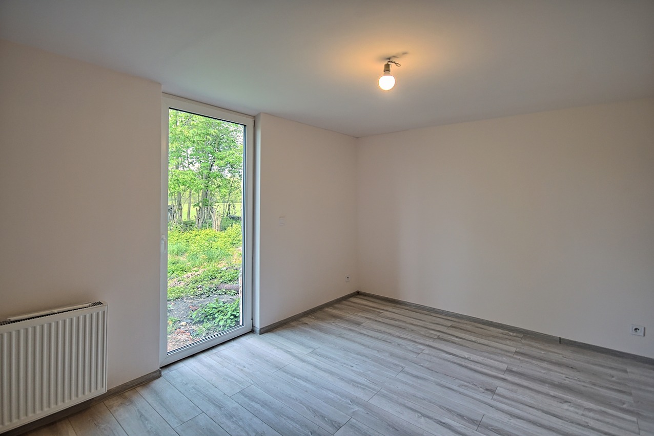 Pusty pokój z nową podłogą imitującą drewno, świeżo pomalowanymi ścianami w odcieniu beżu, grzejnikiem pod oknem i pojedynczą żarówką na suficie.