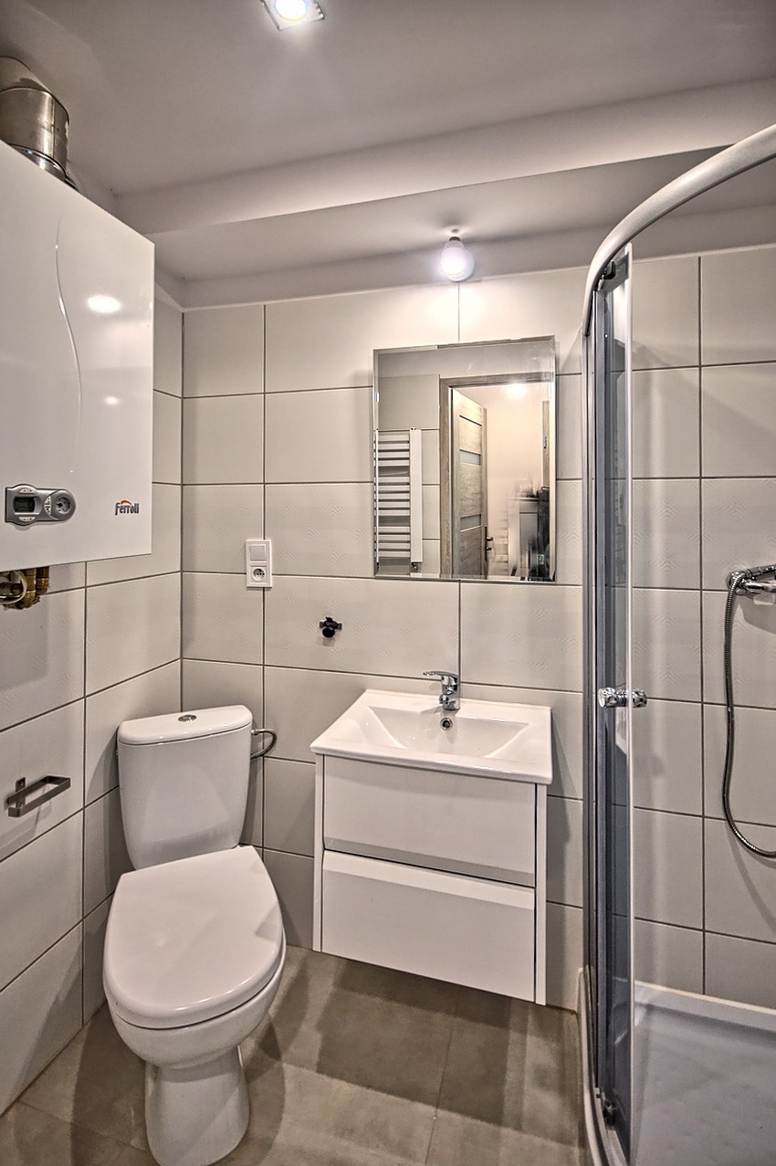 Jasna łazienka z białymi kwadratowymi płytkami, kabiną prysznicową z zaokrąglonymi drzwiami, wiszącą szafką z umywalką, toaletą i piecem gazowym na ścianie.