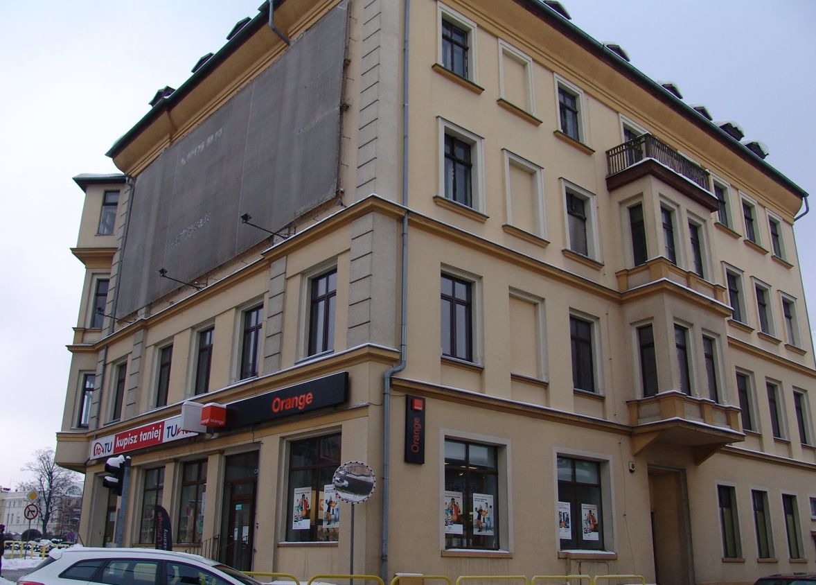 Kremowa kamienica narożna z zasłoniętą reklamą na szczycie, widoczne reklamy Orange na parterze.
