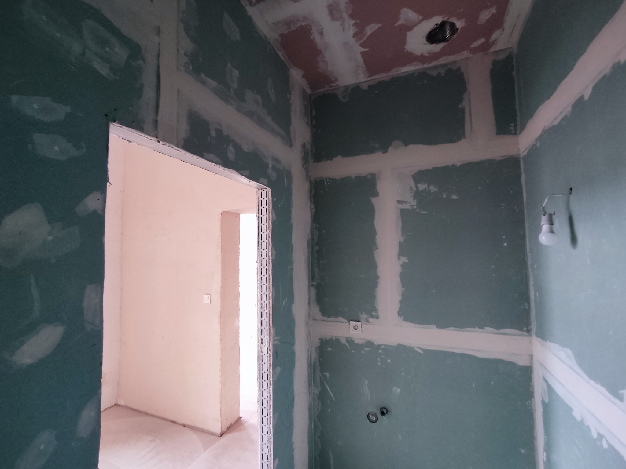 Wnętrze w trakcie prac wykończeniowych: płyty gipsowo-kartonowe na ścianach i suficie, spoinowane łączenia, widoczne puszki elektryczne, surowa ościeżnica drzwiowa.