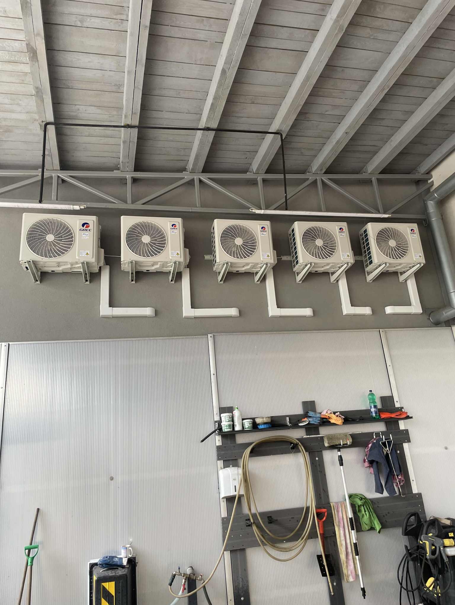 Rząd pięciu klimatyzatorów Gree zamontowanych na szarej ścianie w pomieszczeniu o metalowej konstrukcji sufitu i ścianach z blachy falistej. Widoczne rury odprowadzające.