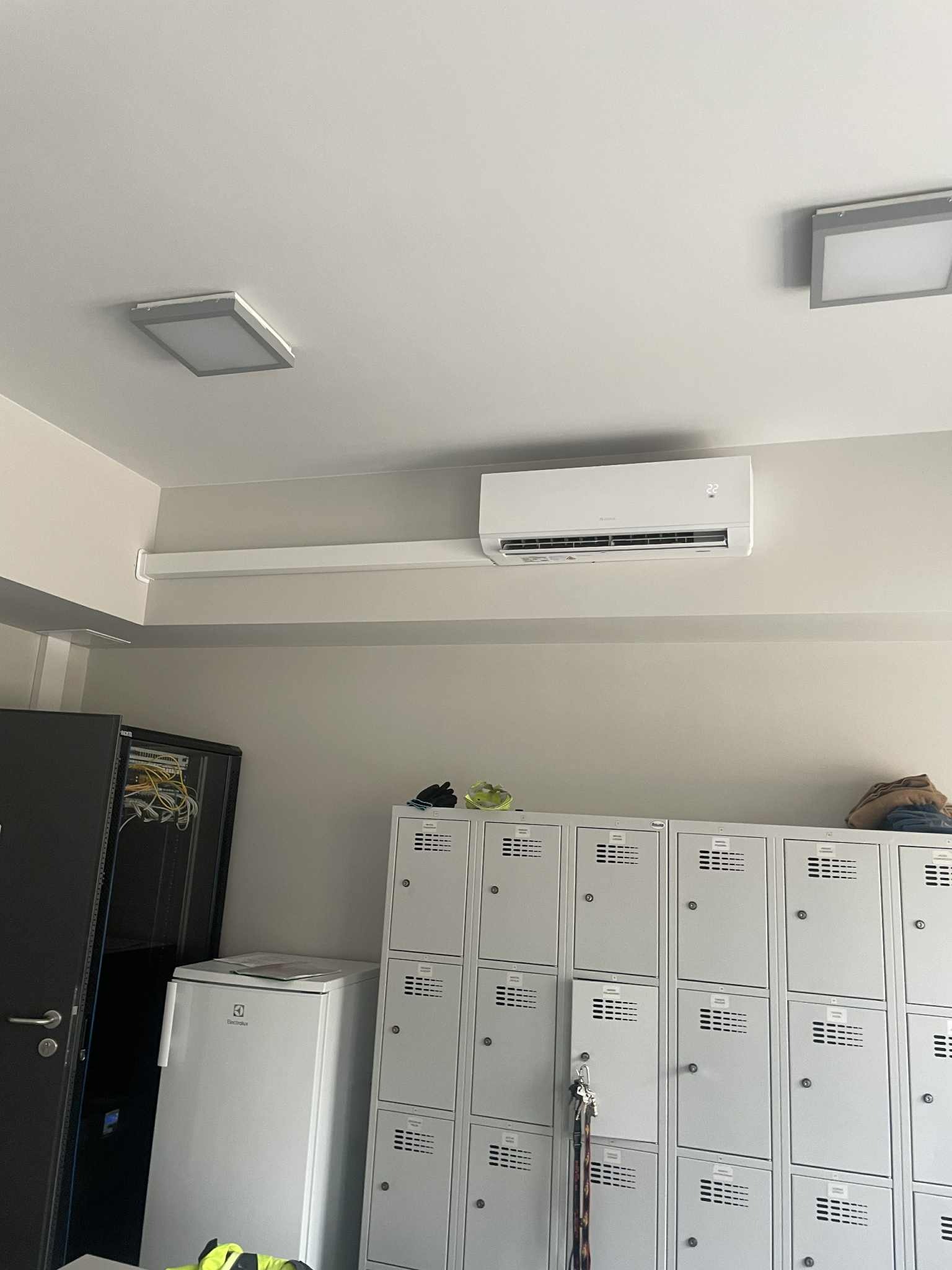 Biały klimatyzator ścienny w pomieszczeniu biurowym z szafkami pracowniczymi, serwerownią i oświetleniem sufitowym. Widok z dołu.