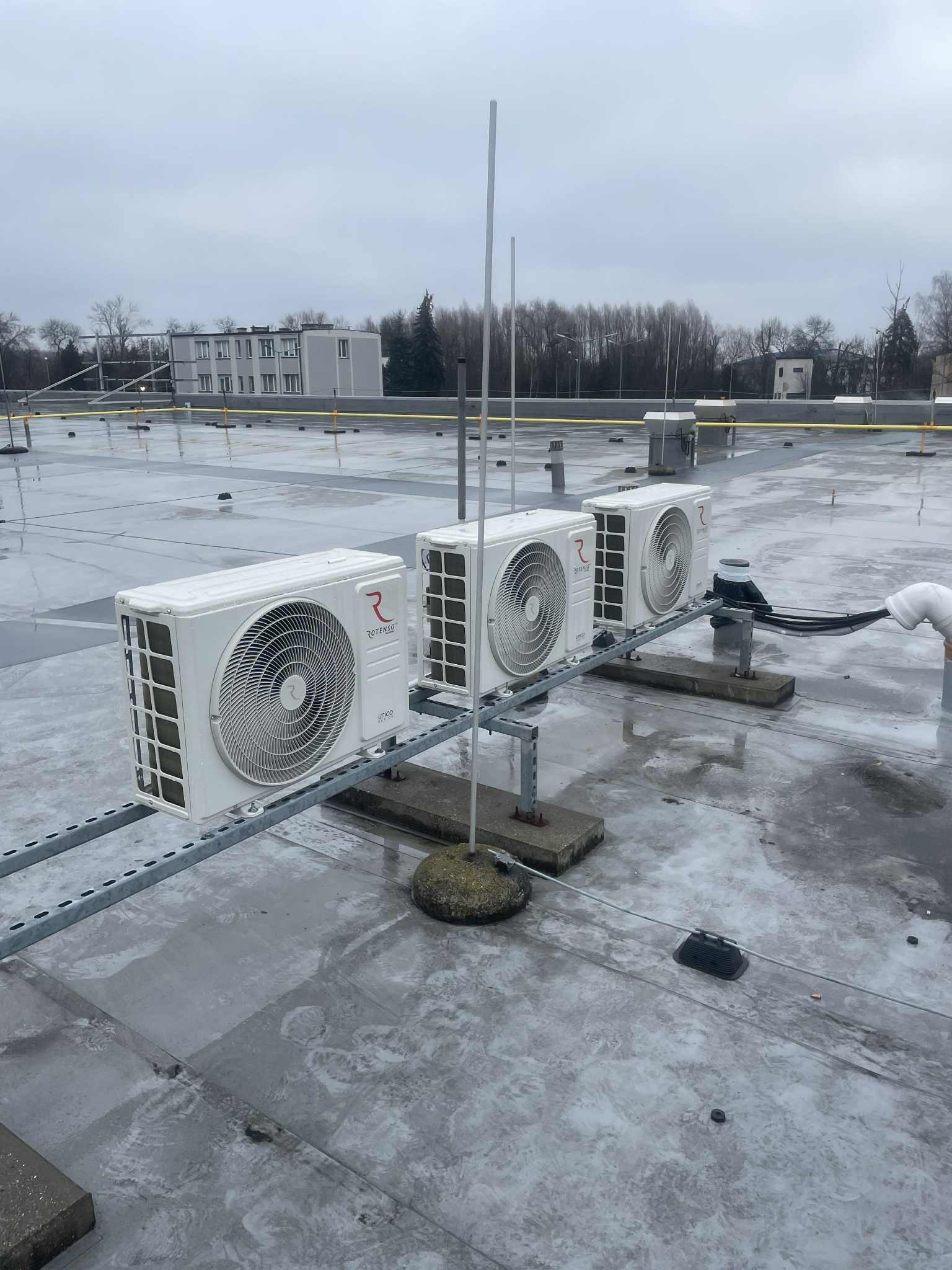 Trzy klimatyzatory Rotenso Unico zamontowane na dachu budynku, widoczne elementy instalacji i mocowania. Szary, pochmurny dzień.