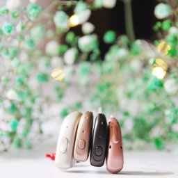 BELL Aparaty Słuchowe Rzeszów - Cztery aparaty słuchowe w różnych odcieniach (beż, brąz, czarny, róż) na białym tle z dekoracją z zielonych kwiatów. Prezentacja nowoczesnych aparatów słuchowych.