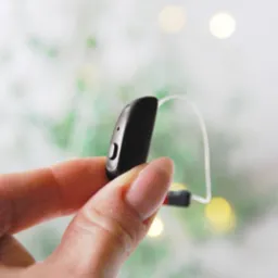 Czarny aparat słuchowy zauszny trzymany w palcach na rozmytym tle z motywem świątecznym.