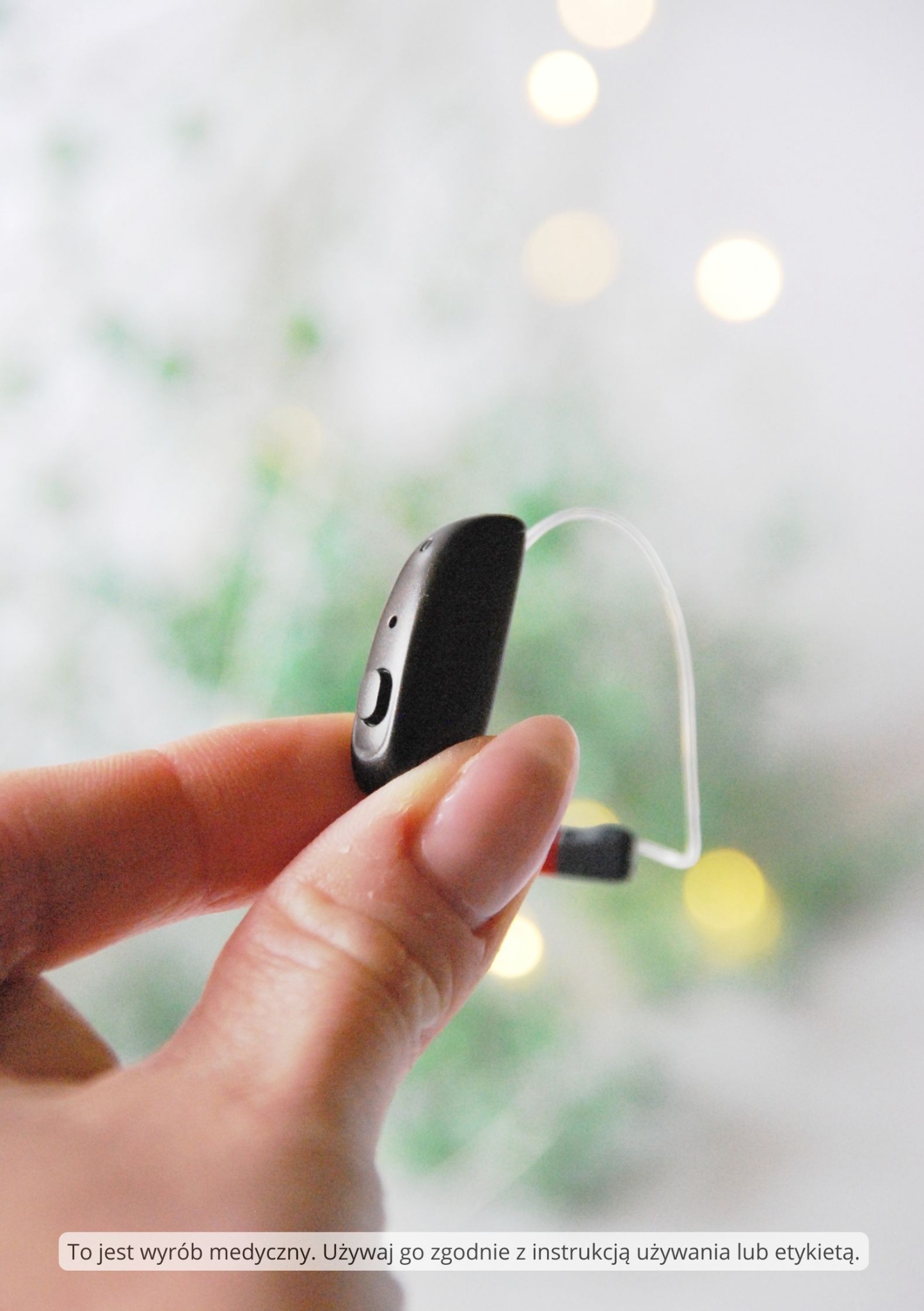 Czarny aparat słuchowy zauszny trzymany w palcach na rozmytym tle z motywem świątecznym.