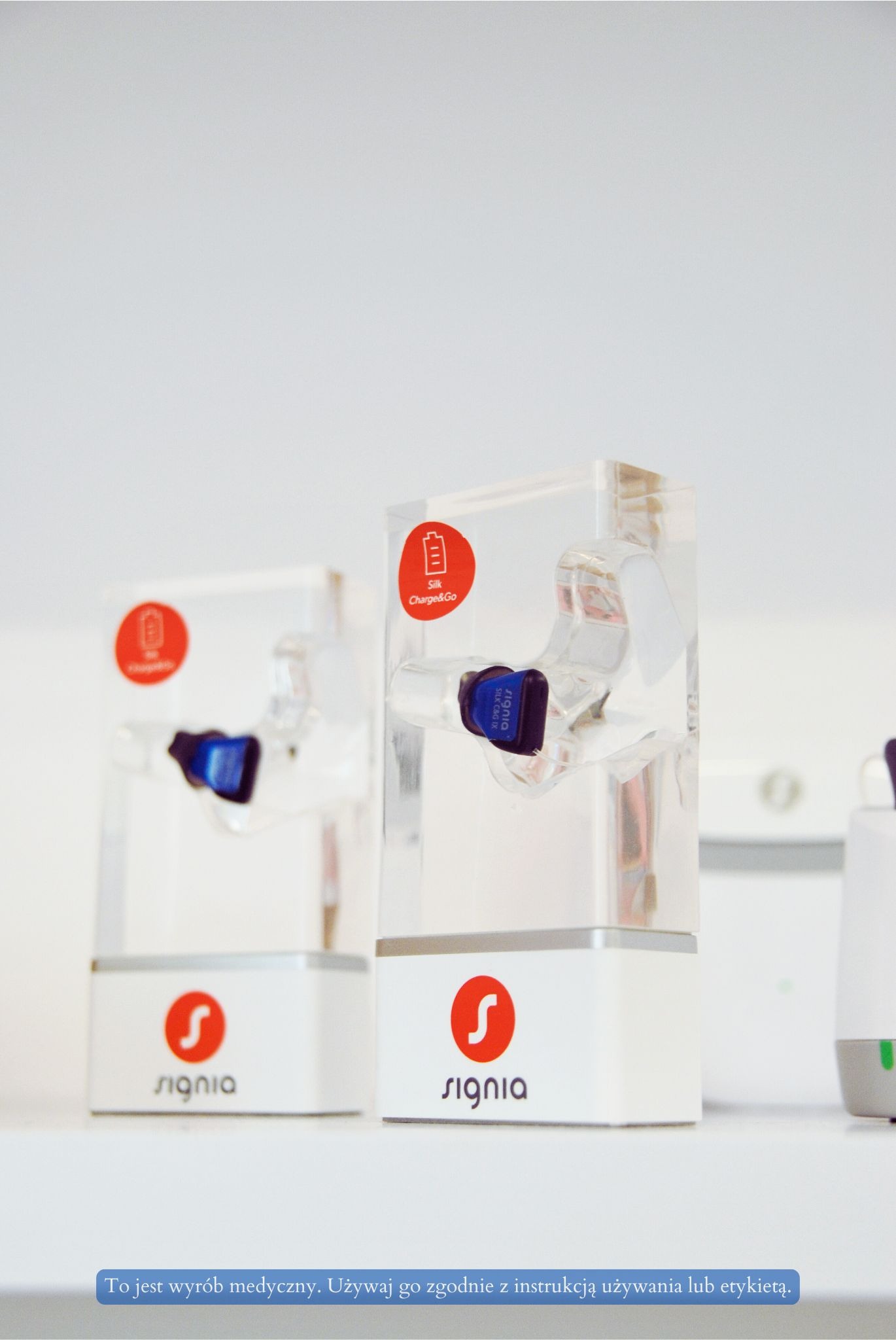 Aparaty słuchowe Signia Silk Charge&Go w przezroczystych stacjach dokujących z czerwonym logo na białym tle. Urządzenia medyczne na jasnej powierzchni.