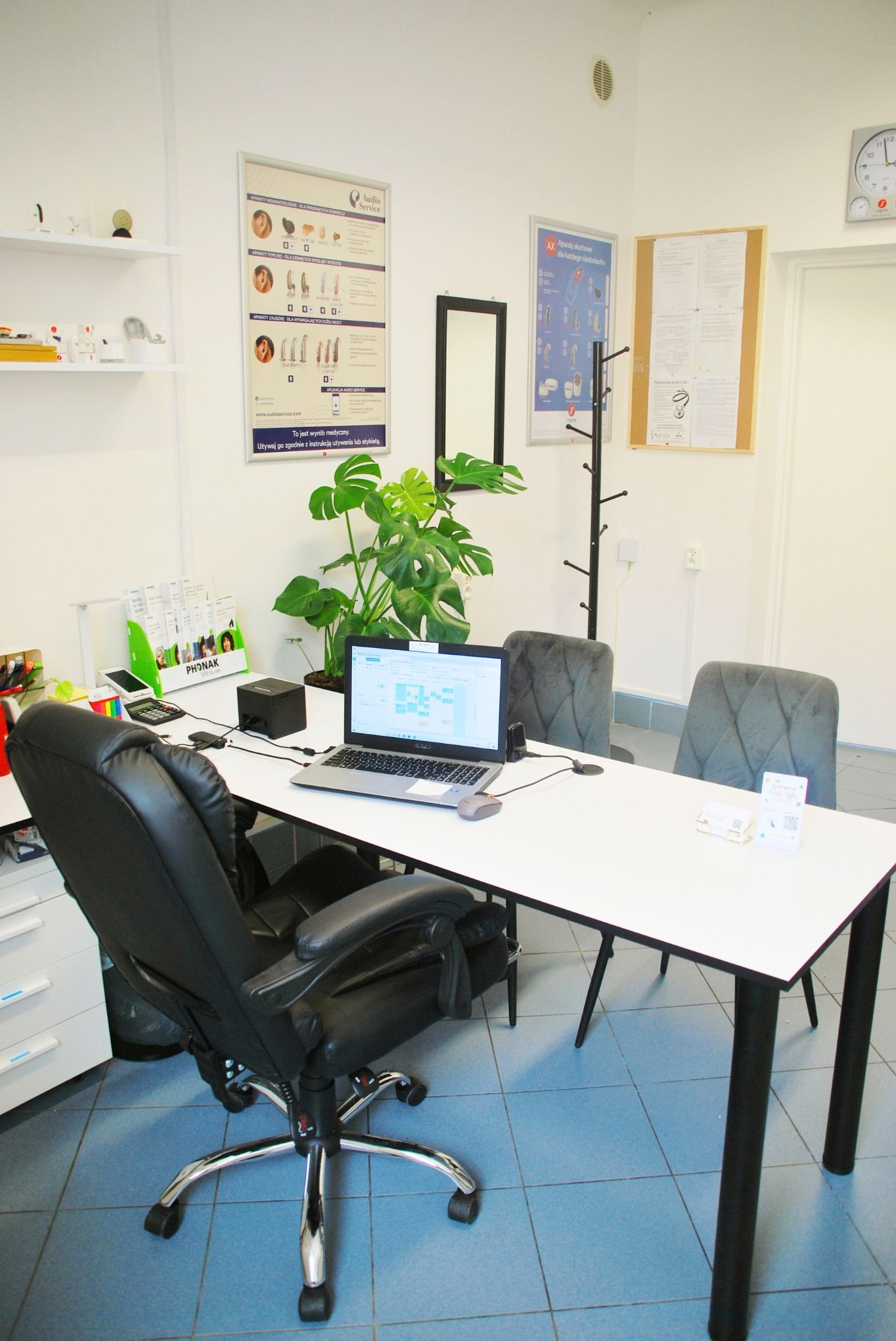 Gabinet lekarski z biurkiem, krzesłami i plakatem o aparatach słuchowych. Na biurku laptop, roślina i materiały informacyjne Phonak.