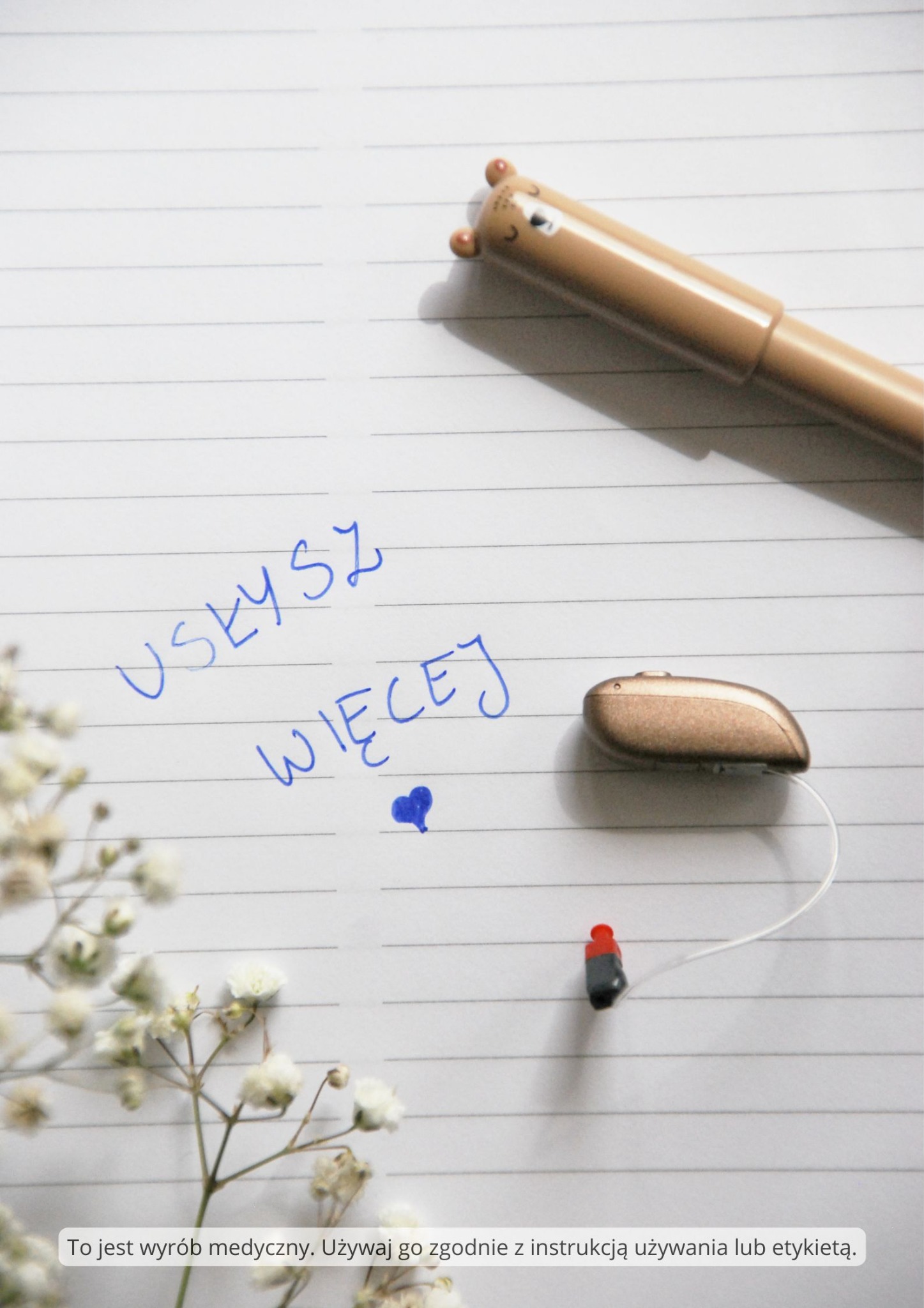 Aparat słuchowy na kartce w linie z napisem 'Usłysz więcej', obok długopis z motywem misia i gałązka gipsówki. Produkt medyczny.