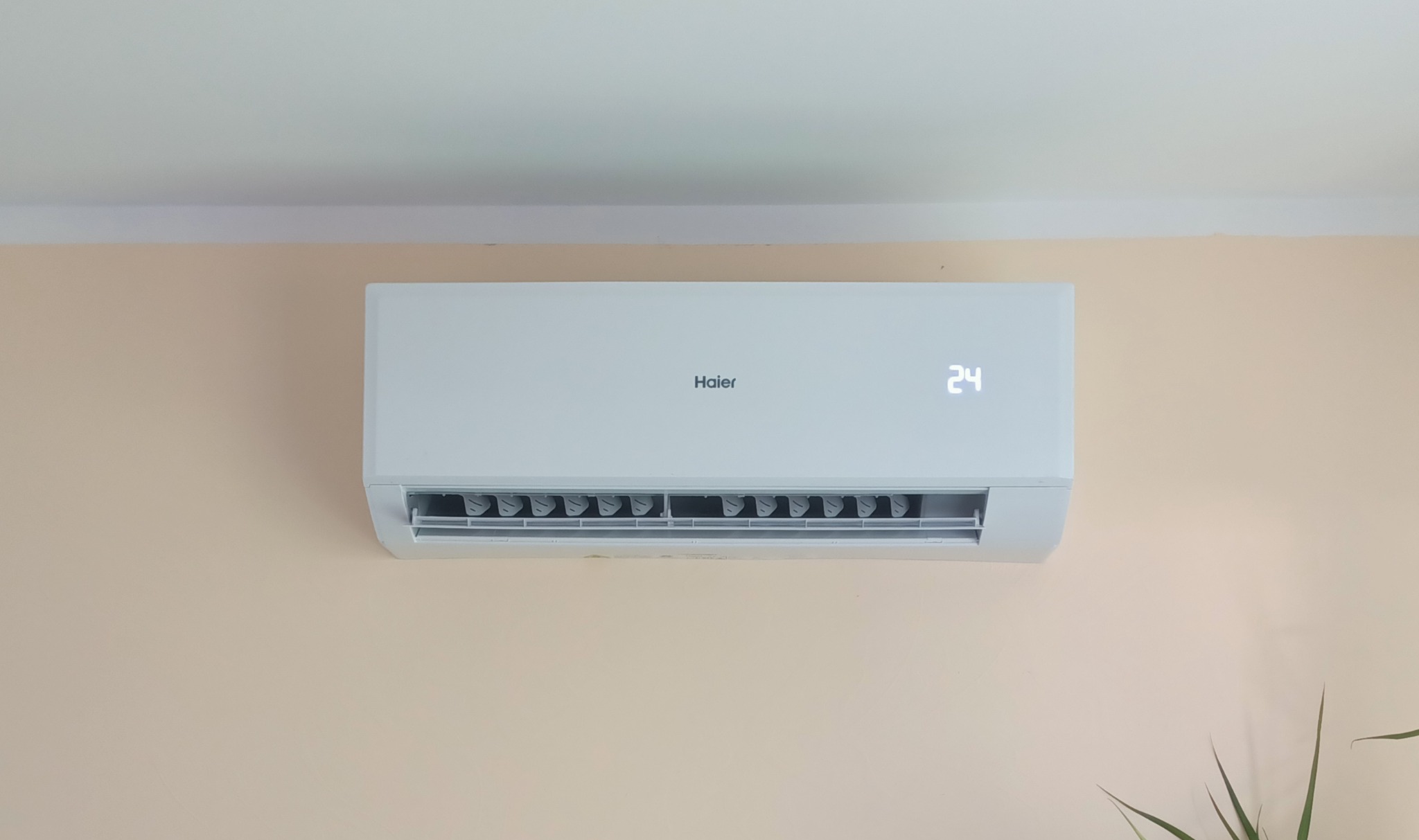 Biały klimatyzator Haier zamontowany na beżowej ścianie, wyświetlający temperaturę 24 stopnie Celsjusza.