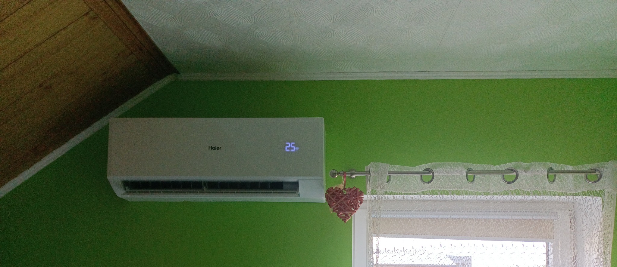 Zamontowany klimatyzator Haier na zielonej ścianie w pokoju z dekoracyjnym sercem wiszącym obok, widoczna temperatura 25 stopni Celsjusza.