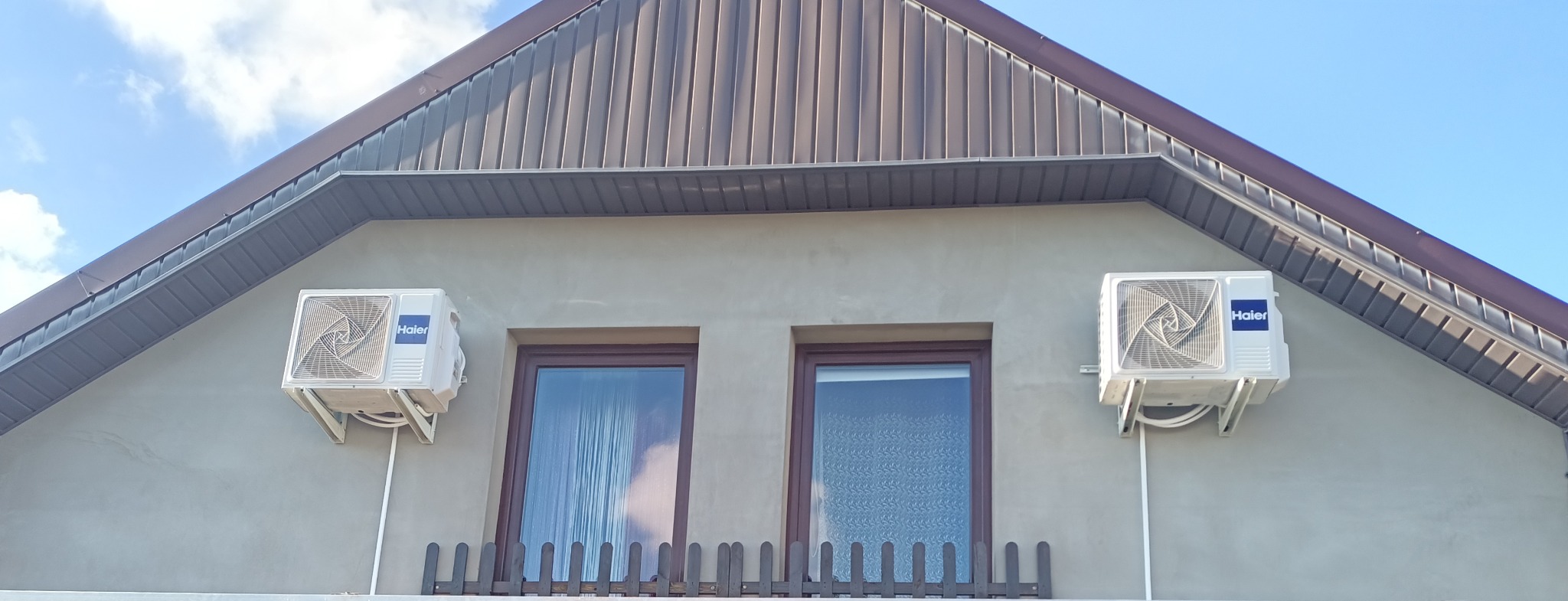 Dwa klimatyzatory Haier zamontowane symetrycznie po bokach okien na elewacji domu z brązowym dachem.
