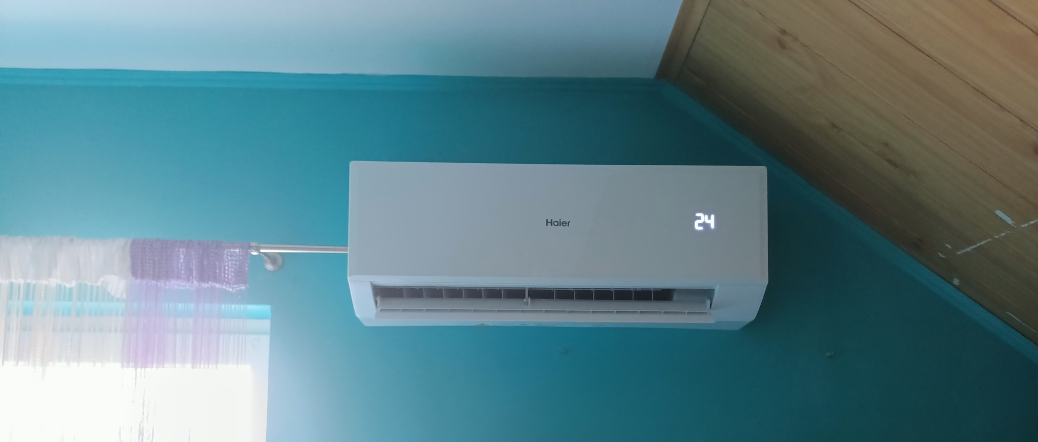 Biała jednostka klimatyzacyjna Haier zamontowana na turkusowej ścianie w pokoju na poddaszu, wyświetlająca temperaturę 24 stopnie Celsjusza.