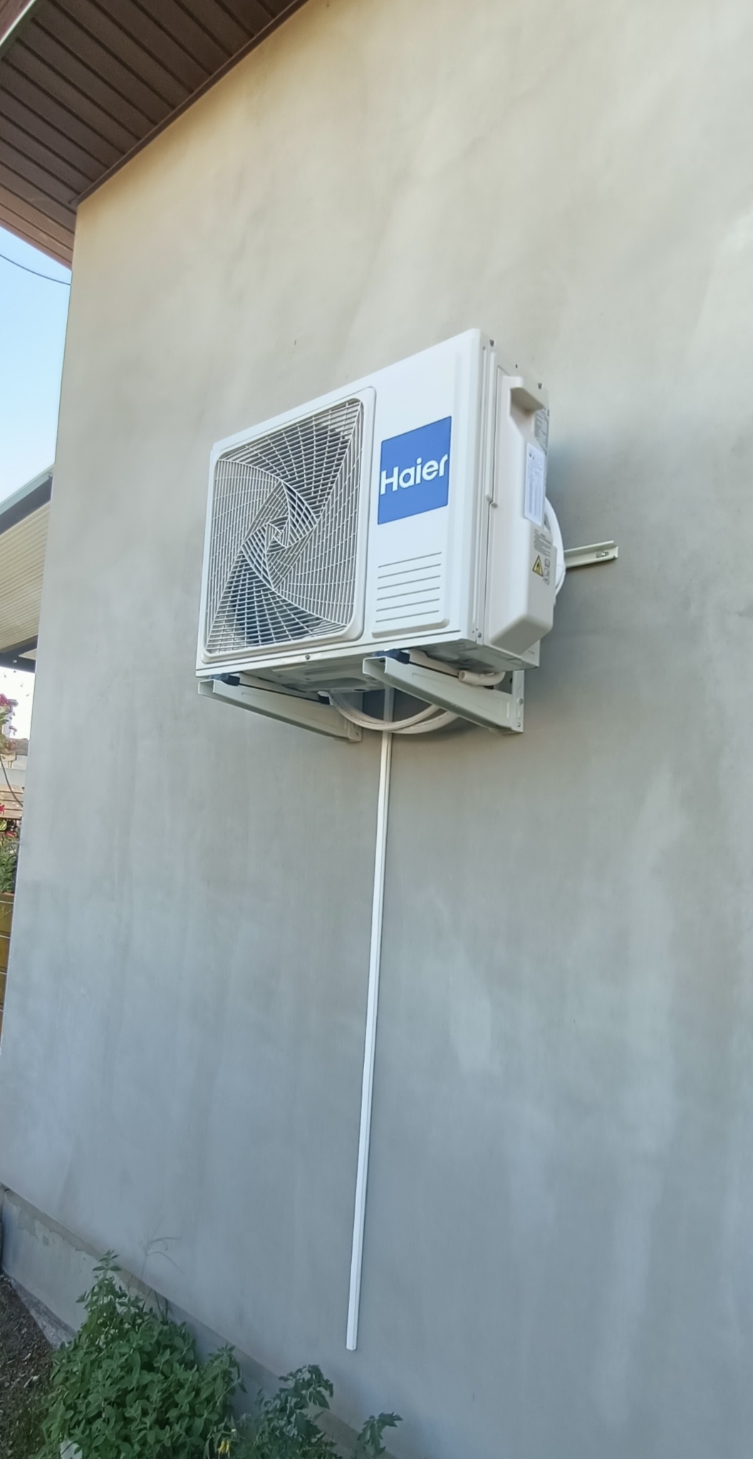 Zewnętrzna jednostka klimatyzacji Haier zamontowana na szarej ścianie budynku, widoczne rury odprowadzające skropliny.