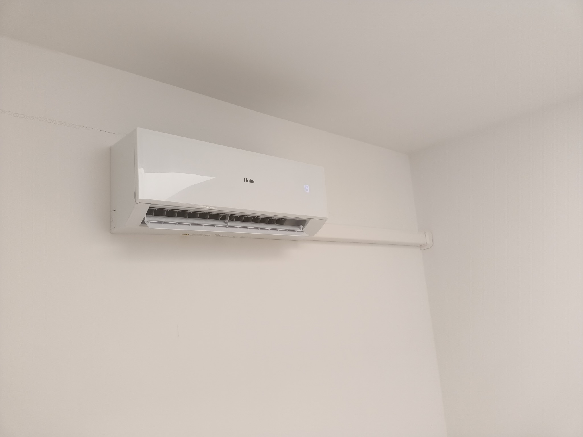 Biała klimatyzacja Haier zamontowana na jasnej ścianie w pomieszczeniu. Widoczny fragment sufitu i rura odprowadzająca skropliny.