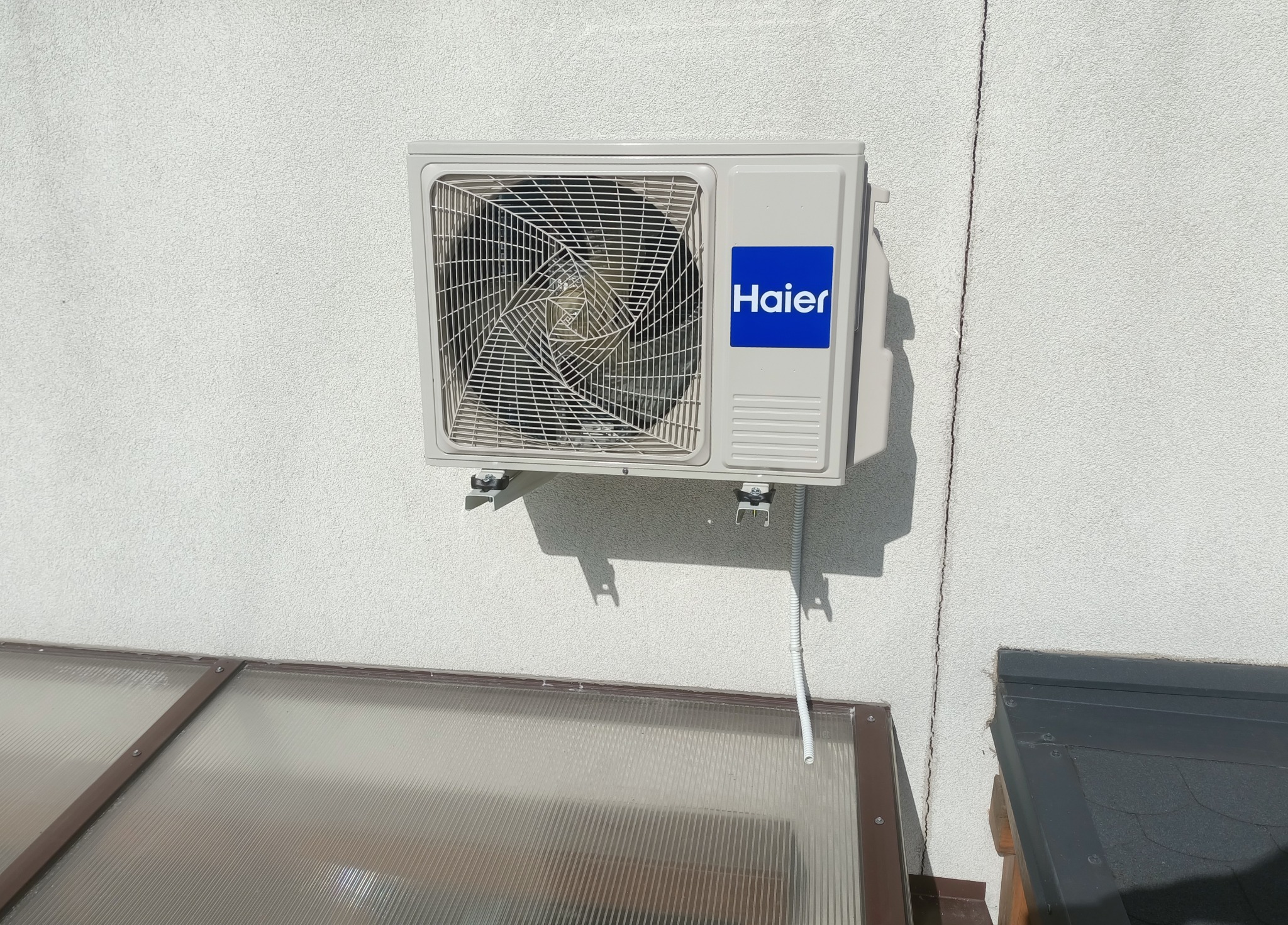 Zewnętrzna jednostka klimatyzacji Haier zamontowana na szarej, chropowatej ścianie budynku. Widoczny fragment dachu i oranżerii.