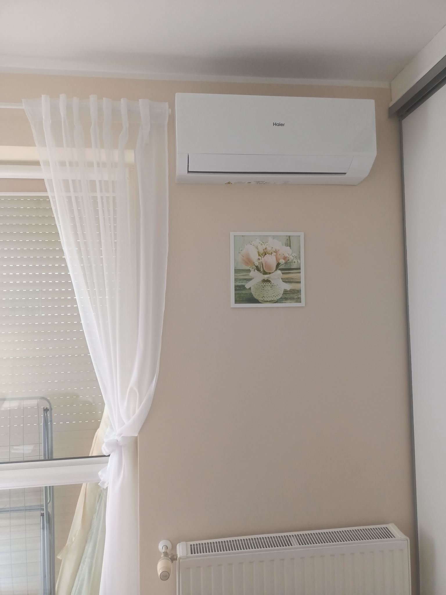 Biała klimatyzacja Haier na ścianie w jasnym pokoju, nad obrazkiem z kwiatami. Obok okno z firanką i grzejnik. Widok z dołu, neutralne oświetlenie.