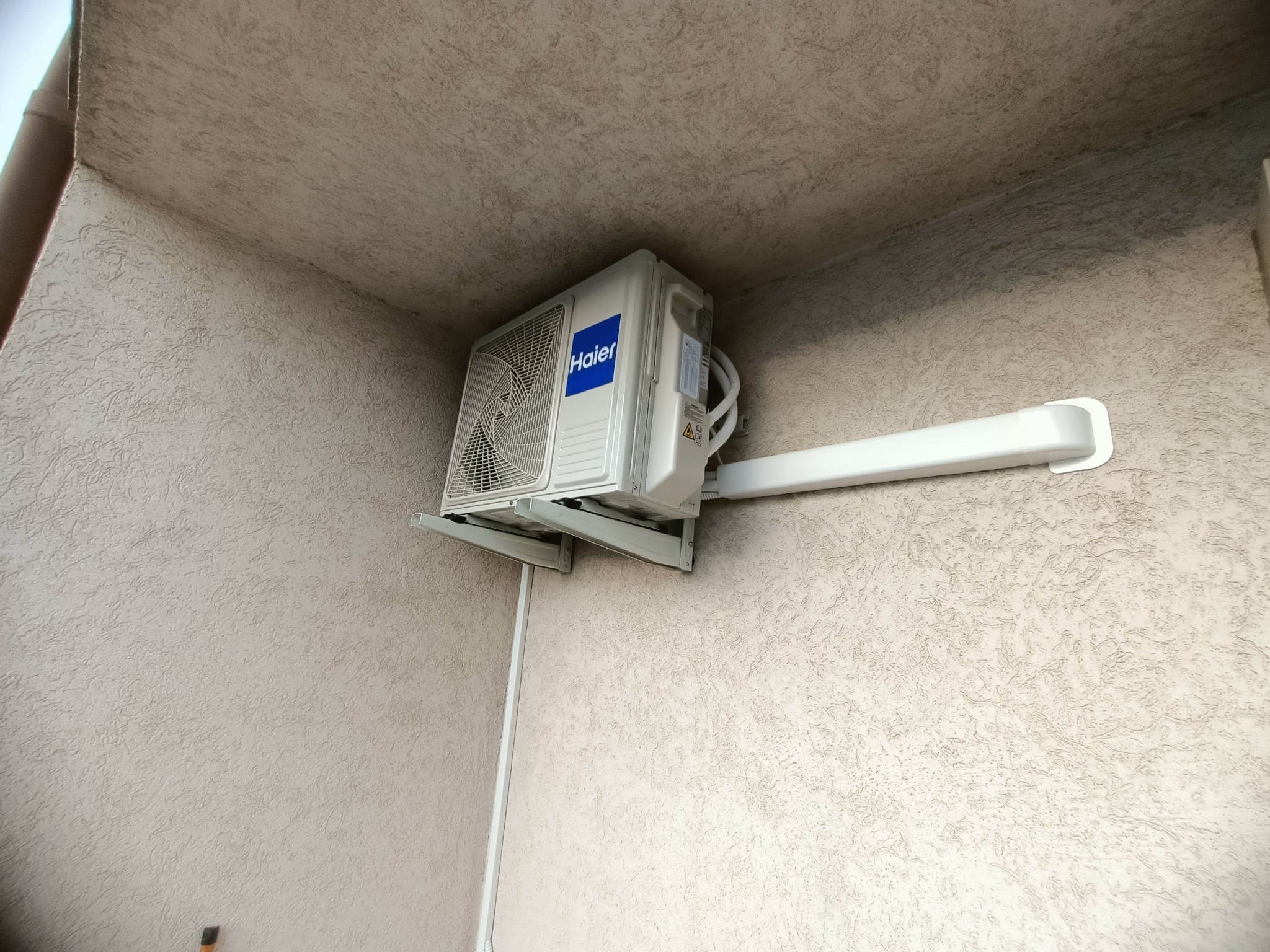 Klimatyzator Haier zamontowany na zewnątrz budynku, widoczny z dołu, z przymocowanymi rurami i wspornikiem. Teksturowana ściana w tle.