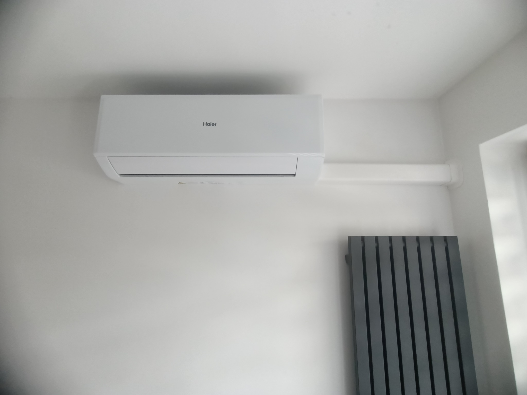 Biały klimatyzator Haier zamontowany na ścianie w jasnym pomieszczeniu, widoczny fragment grzejnika. Instalacja poprowadzona wzdłuż ściany.