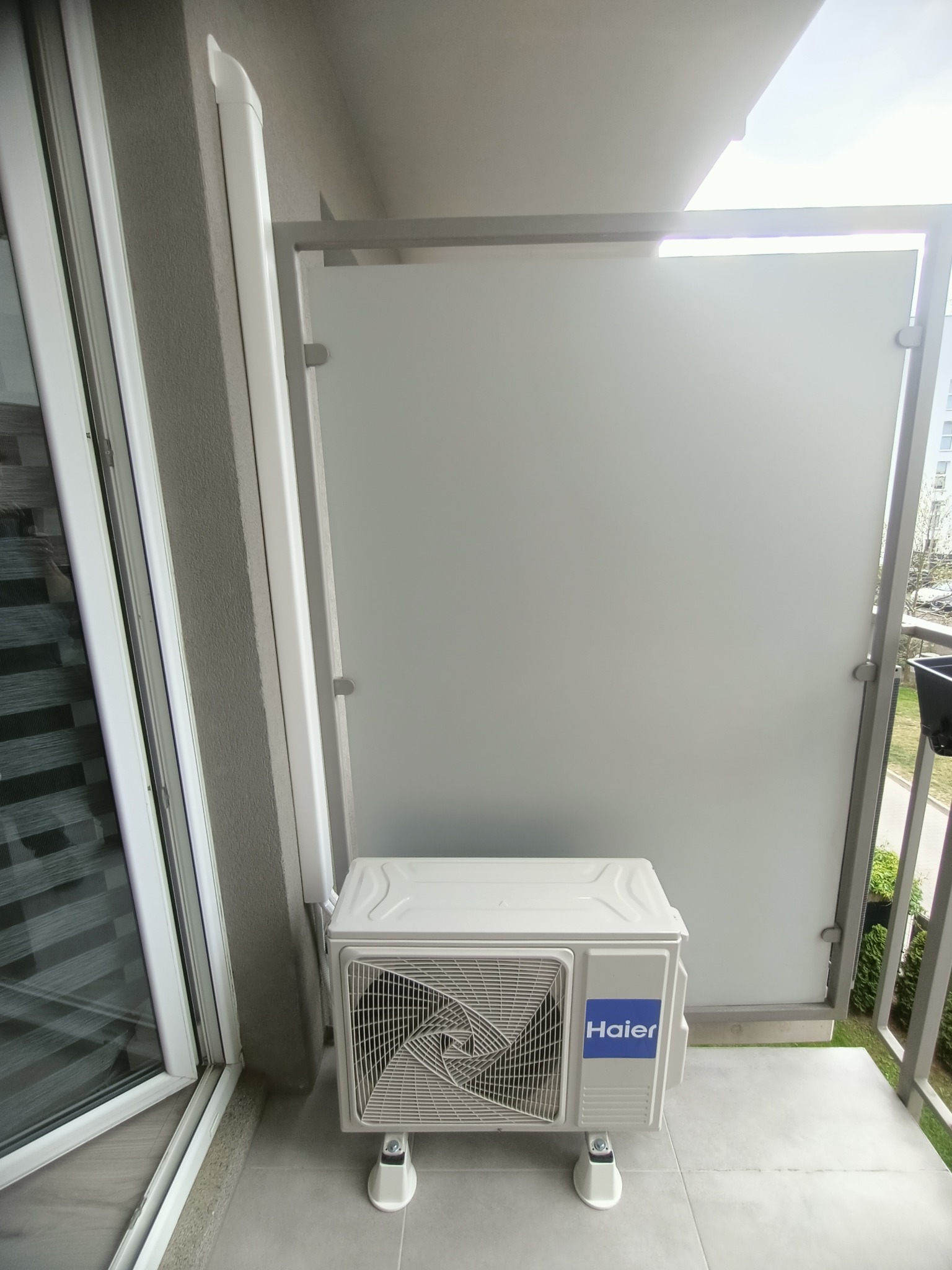 Biała jednostka zewnętrzna klimatyzacji Haier na balkonie z szarą podłogą i szklaną balustradą. Widoczne elementy instalacji przy ścianie.