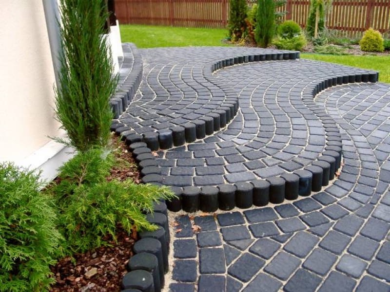 Czarna kostka brukowa ułożona w falujący wzór z obrzeżami z drewnianych palików, otoczona zielenią i niskimi krzewami.