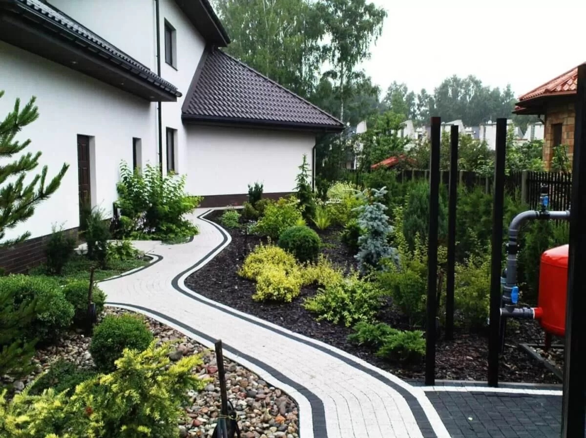 Kręta ścieżka z jasnej kostki brukowej z czarnym obramowaniem, wijąca się przez zadbany ogród z różnorodną roślinnością, widok na biały dom z brązowym dachem w tle.