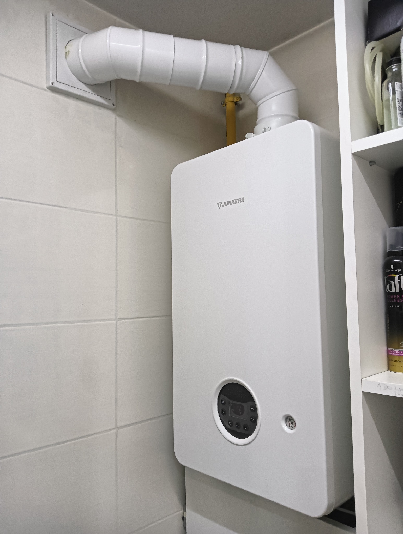 Nowy, biały piec gazowy marki Junkers zamontowany na ścianie wyłożonej jasnymi płytkami, z widocznymi białymi rurami odprowadzającymi spaliny i żółtą rurą gazową, obok półki z kosmetykami.