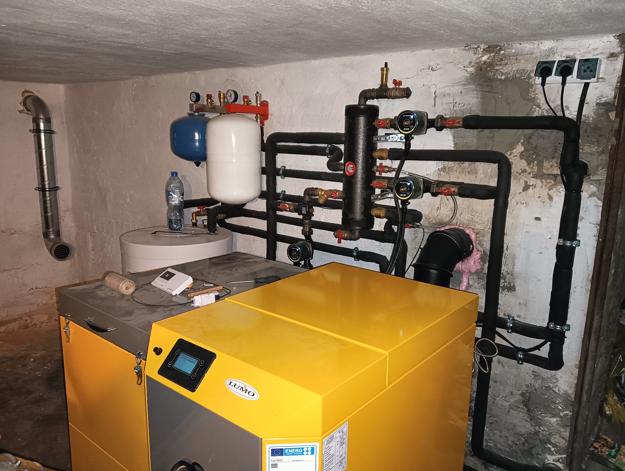 Żółty piec grzewczy LUMO z podłączonymi czarnymi rurami instalacji, manometry, zbiorniki wyrównawcze, butelka wody na zbiorniku, widok w kotłowni z surowymi ścianami.