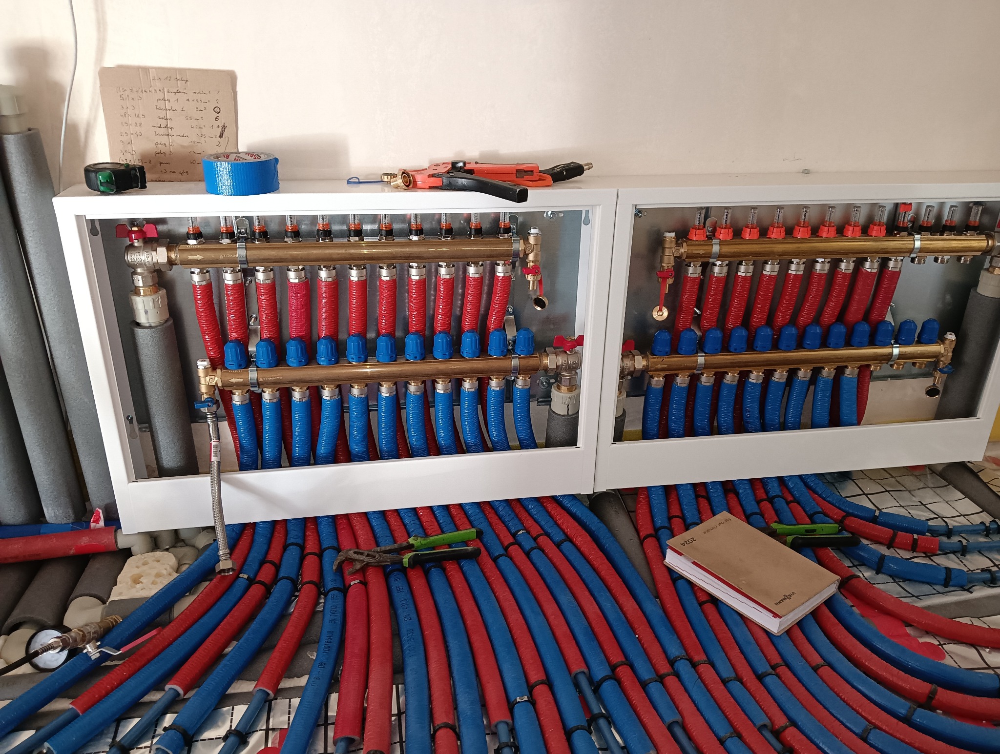 Rozdzielacz ogrzewania podłogowego w białej szafce, z widocznymi czerwono-niebieskimi rurami, narzędziami i notatnikiem Viessmann leżącym na podłodze z siatką montażową.