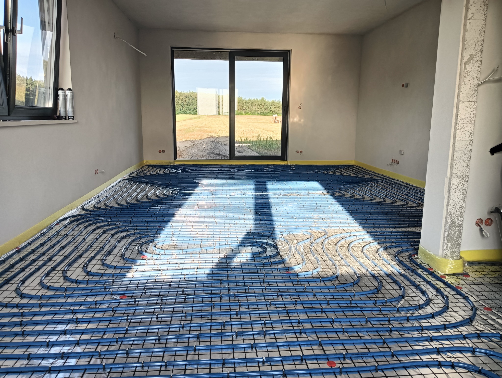 Niebieskie rury ogrzewania podłogowego ułożone na czarnej siatce w surowym wnętrzu z widokiem na pole przez duże okno przesuwne.