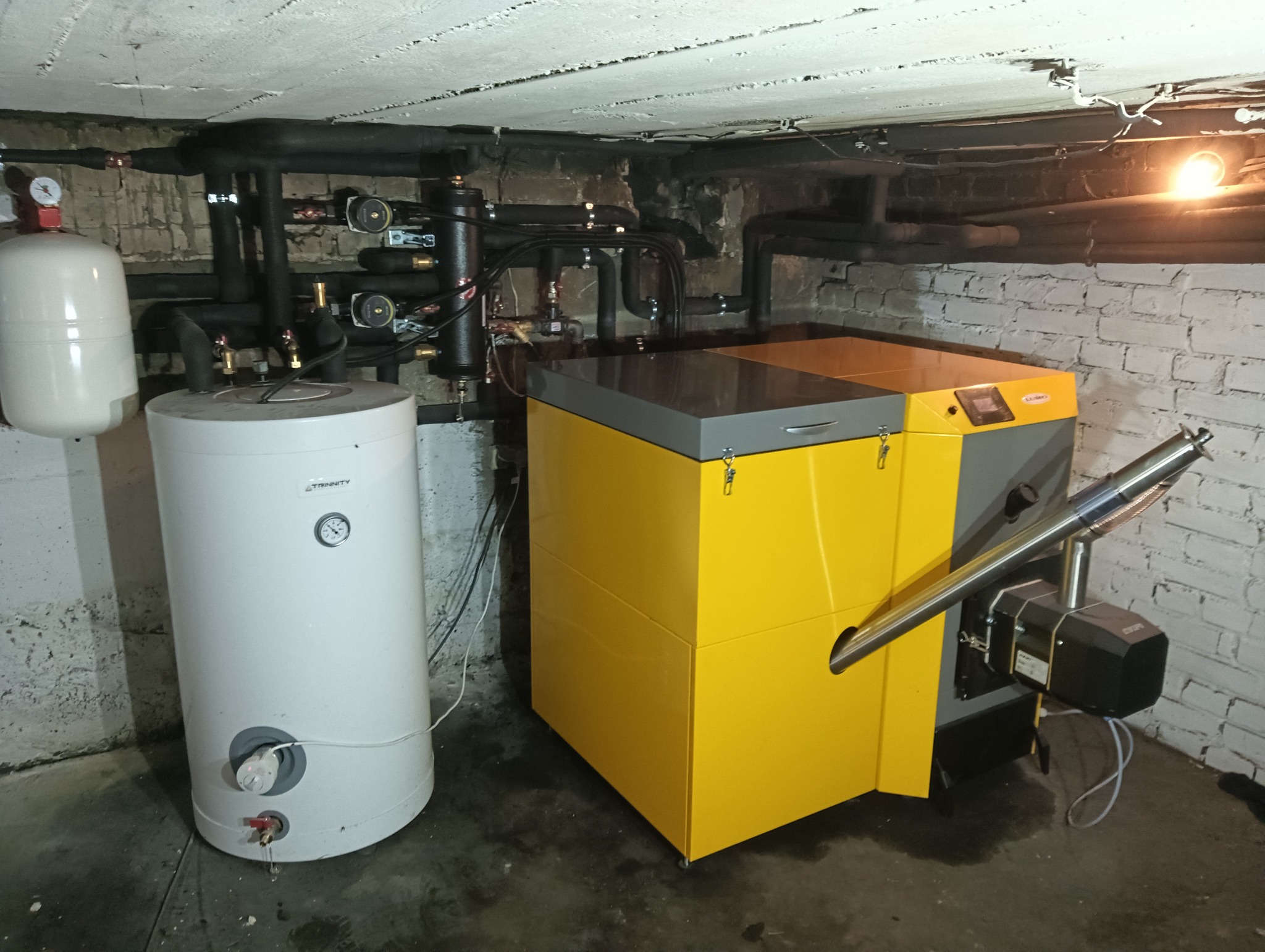 Żółto-szary piec grzewczy z podajnikiem, obok biały zasobnik ciepłej wody, widoczne rury instalacji grzewczej pod sufitem piwnicy.