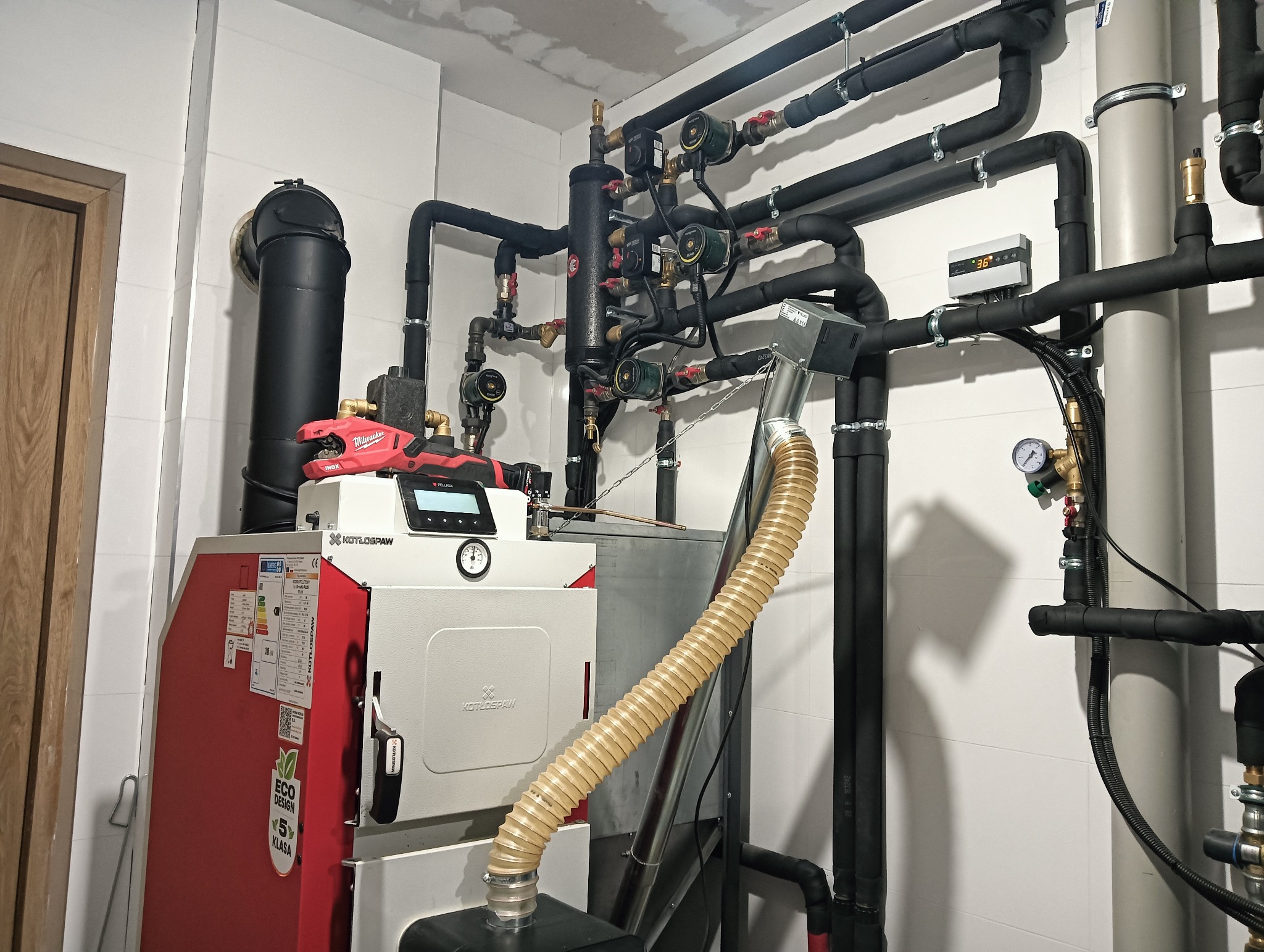 Złożona instalacja kotła na paliwo stałe z widocznym kotłem z podajnikiem, elementami hydraulicznymi, rurami izolowanymi termicznie i narzędziem do cięcia rur na górze kotła.