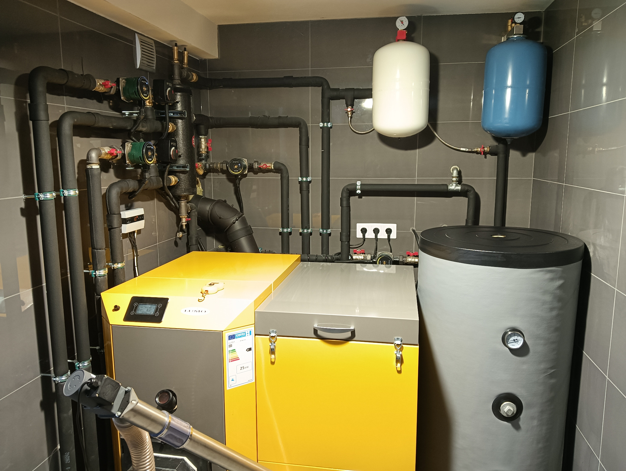 Kotłownia z nowoczesnym piecem dwufunkcyjnym, zbiornikiem akumulacyjnym i rozbudowaną instalacją hydrauliczną na tle szarych płytek.