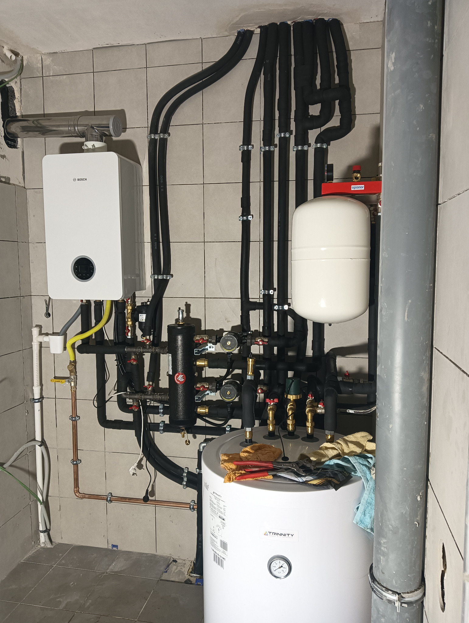Złożona instalacja grzewcza w kotłowni z piecem gazowym Bosch, zasobnikiem Trinity, czarnymi rurami izolowanymi i licznymi zaworami, widok na płytki ceramiczne na ścianie i narzędzia na zasobniku.