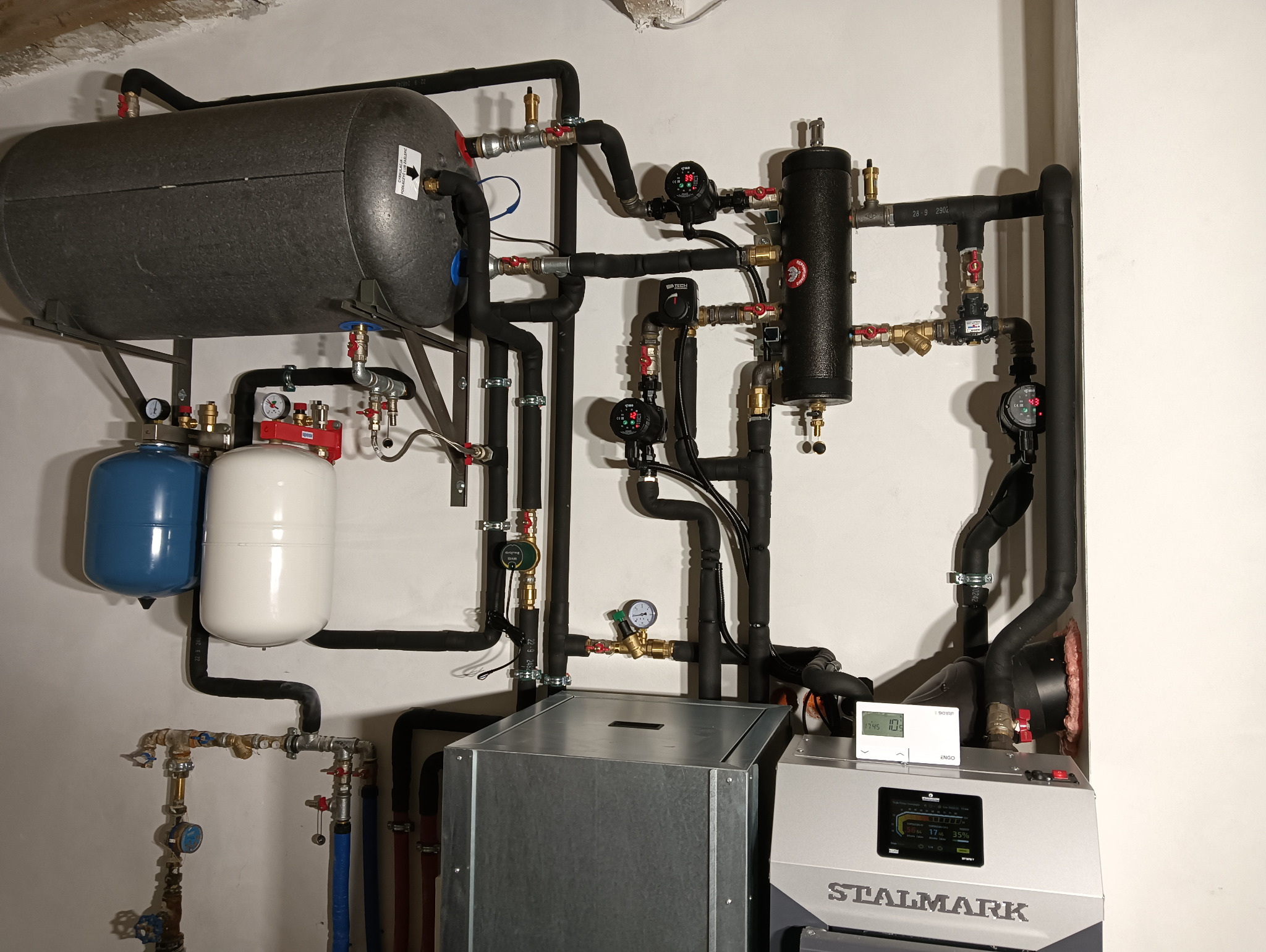 Złożona instalacja grzewcza z piecem Stalmark, zbiornikiem akumulacyjnym, dwoma zbiornikami przeponowymi (niebieski i biały), licznymi rurami, zaworami i manometrami na ścianie.