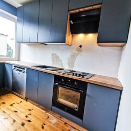 WOJCIECH GRĘBOWICZ - Nowoczesna kuchnia z szarymi szafkami, drewnianym blatem i czarnym sprzętem AGD, w tym piekarnikiem i płytą gazową. Widoczne ślady po remoncie na ścianie i podłodze.