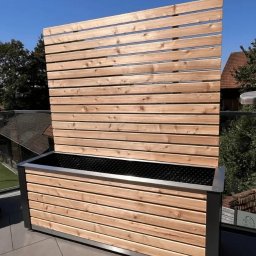 WOJCIECH GRĘBOWICZ - Nowoczesna drewniana donica z panelem osłonowym, wykonana z jasnego drewna z metalowymi akcentami, na tarasie z widokiem na dom i błękitne niebo.