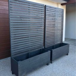 WOJCIECH GRĘBOWICZ - Dwa ciemnoszare, drewniane panele z poziomymi listwami, połączone z donicami, stoją na żwirowym podłożu przed jasną ścianą budynku i brązową bramą garażową.