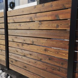 WOJCIECH GRĘBOWICZ - Nowoczesne ogrodzenie z poziomo ułożonych, impregnowanych desek z widocznym usłojeniem, czarne metalowe słupki, śnieg u podstawy, budynek w tle.