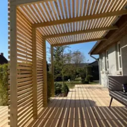 Drewniana konstrukcja z listew tworząca zacienione przejście na tarasie, widok na dom i ogród w słoneczny dzień.
